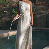 A woman stands barefoot by the pool, surrounded by greenery and flowers, wearing the AYA Sacred Wear Greek Goddess Dress—an organic cotton, white boho dress with a pleated one-shoulder design and braided belt. Sunlight casts soft shadows.