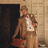 A man wearing a Brown Linen Blazer Jacket for Men from AYA Sacred Wear leans casually against a wooden chair in a sunlit, rustic setting. The background consists of a wooden door and a textured wall adorned with decorative patterns, as sunlight streams through, creating gentle shadows.
