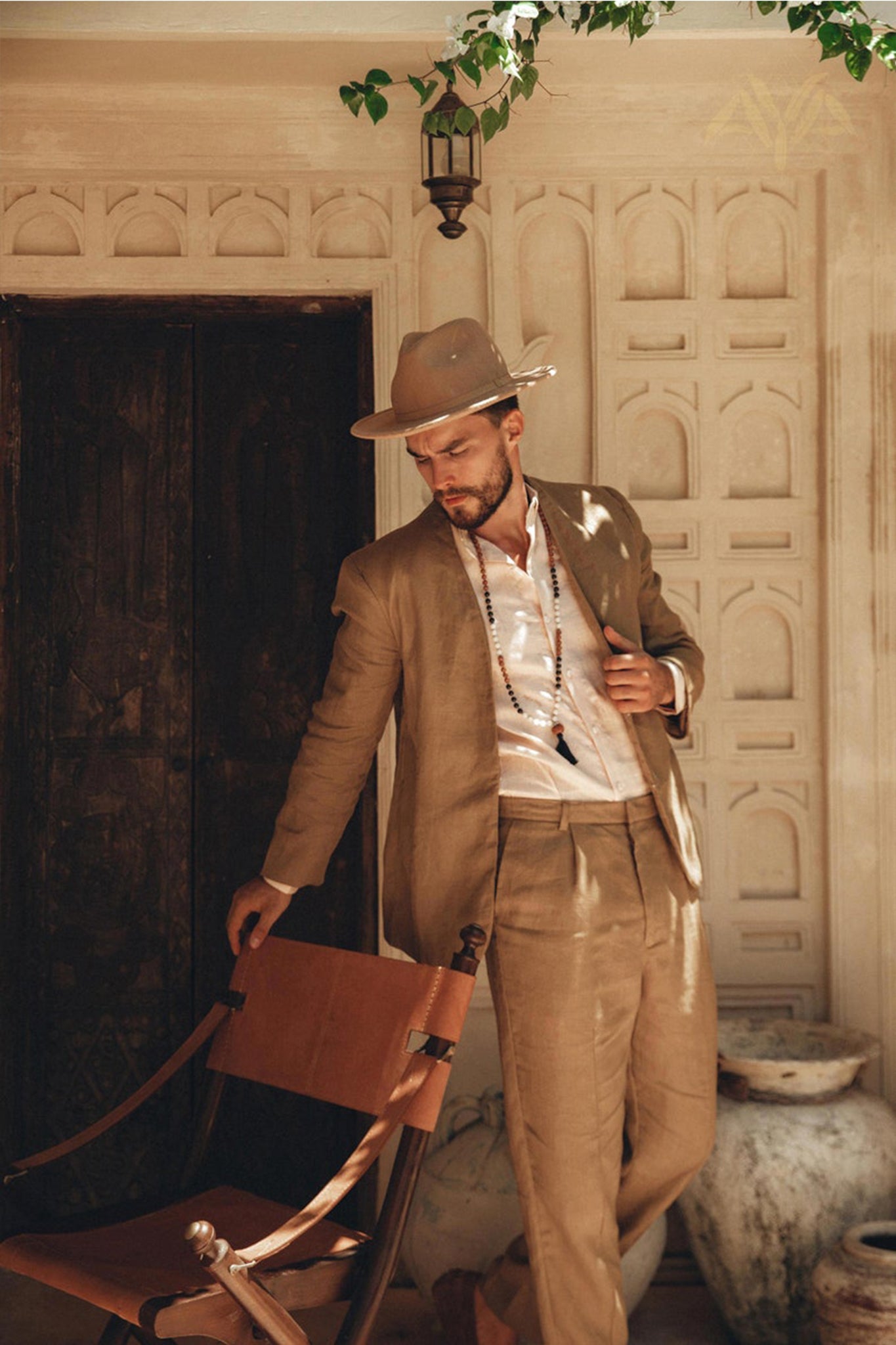 A man wearing a Brown Linen Blazer Jacket for Men from AYA Sacred Wear leans casually against a wooden chair in a sunlit, rustic setting. The background consists of a wooden door and a textured wall adorned with decorative patterns, as sunlight streams through, creating gentle shadows.