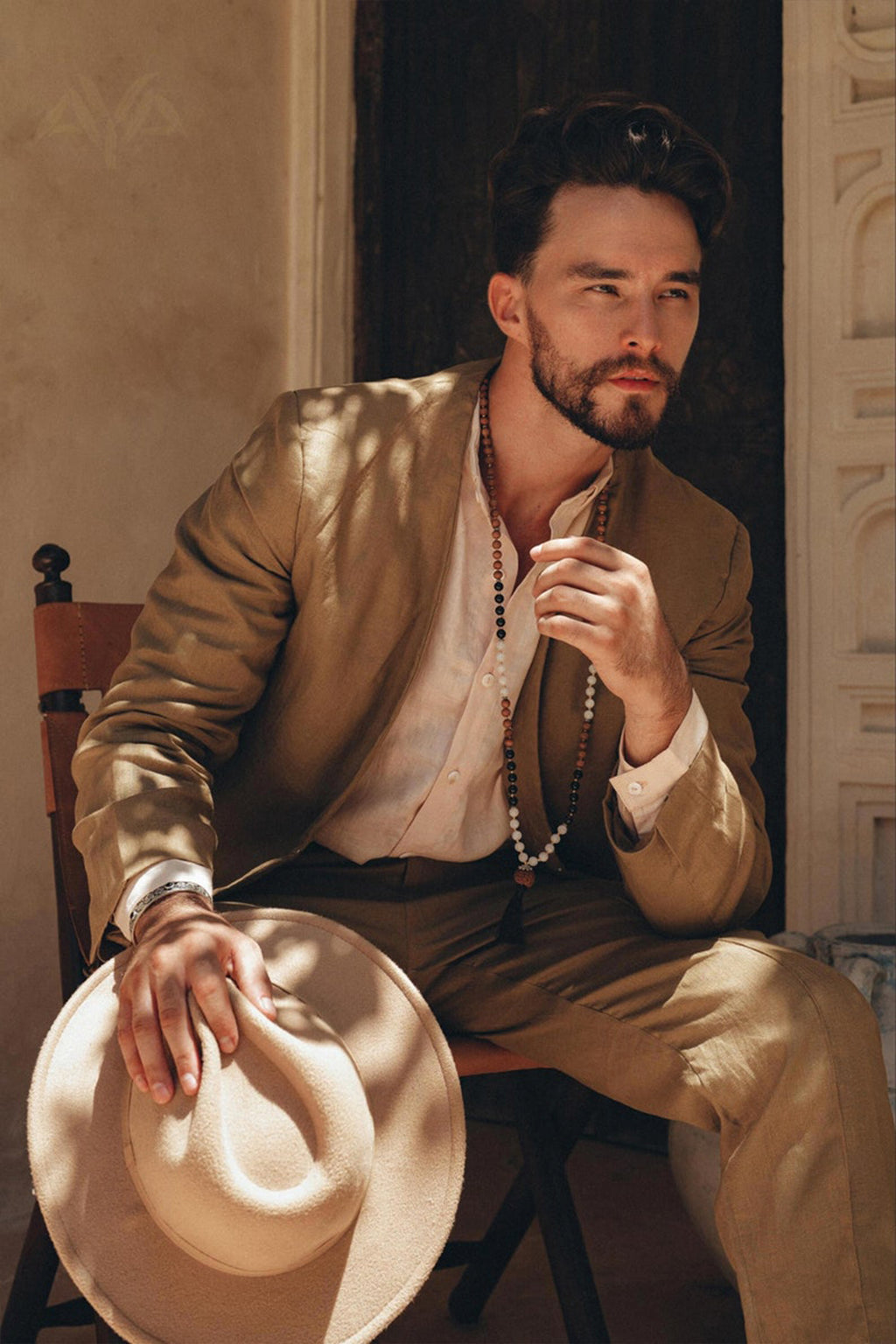 A bearded individual sits on a chair, wearing the AYA Sacred Wear Brown Linen Blazer Jacket for Men, botanically dyed in light brown. They hold a matching hat and appear thoughtful as sunlight creates soft shadows on their attire. The backdrop showcases a textured wall with a partially open door.