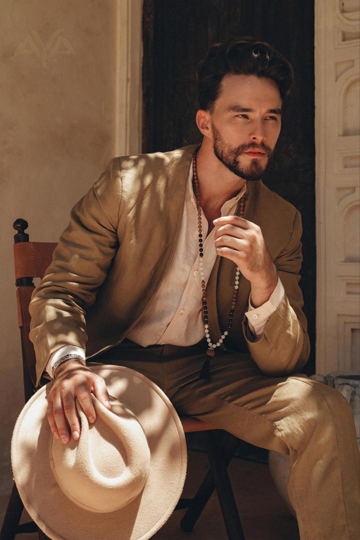 A bearded individual sits on a chair, wearing the AYA Sacred Wear Brown Linen Blazer Jacket for Men, botanically dyed in light brown. They hold a matching hat and appear thoughtful as sunlight creates soft shadows on their attire. The backdrop showcases a textured wall with a partially open door.