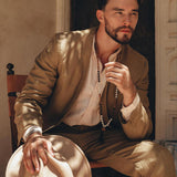 A bearded individual sits on a chair, wearing the AYA Sacred Wear Brown Linen Blazer Jacket for Men, botanically dyed in light brown. They hold a matching hat and appear thoughtful as sunlight creates soft shadows on their attire. The backdrop showcases a textured wall with a partially open door.