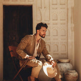 A man with a beard sits on a wooden chair outdoors, wearing the Brown Linen Blazer Jacket for Men by AYA Sacred Wear and holding a hat. The background showcases textured white walls and large clay pots, with sunlight casting soft shadows to create a warm and serene atmosphere.
