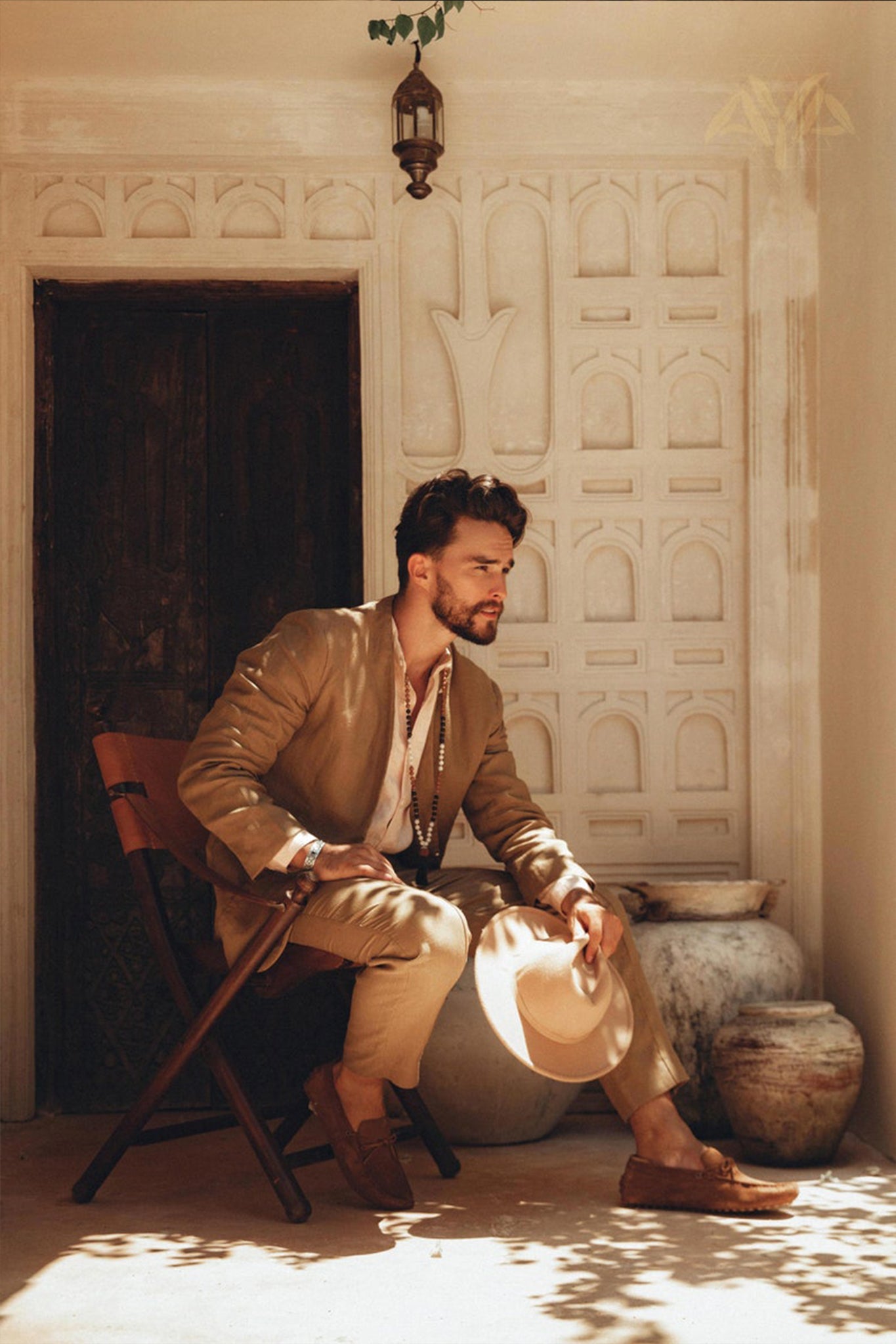 A man with a beard sits on a wooden chair outdoors, wearing the Brown Linen Blazer Jacket for Men by AYA Sacred Wear and holding a hat. The background showcases textured white walls and large clay pots, with sunlight casting soft shadows to create a warm and serene atmosphere.