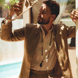 A man in an AYA Sacred Wear Brown Linen Blazer Jacket holds a hat and gazes at the white flowers above him. He's outdoors, with sunlight casting intricate patterns of light and shadow on his outfit, while a swimming pool can be seen in the background.