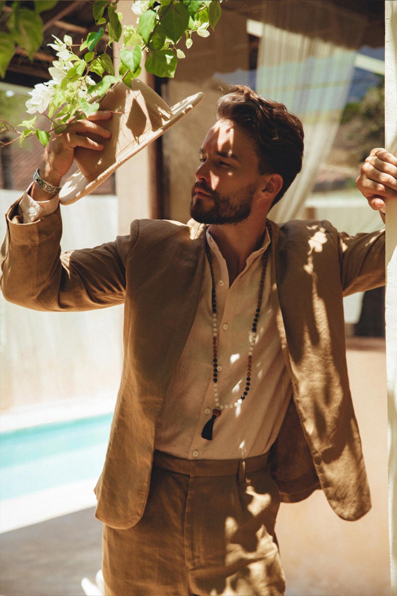 A man in an AYA Sacred Wear Brown Linen Blazer Jacket holds a hat and gazes at the white flowers above him. He's outdoors, with sunlight casting intricate patterns of light and shadow on his outfit, while a swimming pool can be seen in the background.