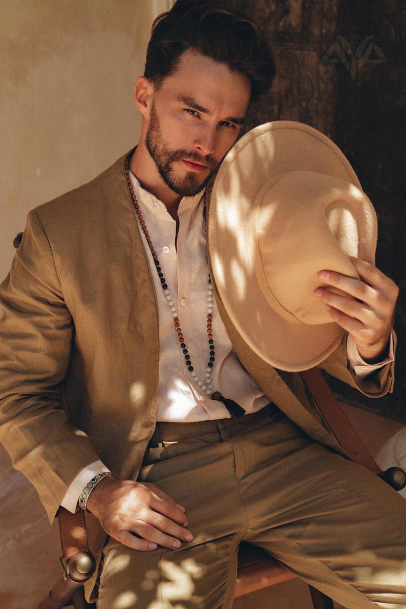 A man sits on a chair wearing an AYA Sacred Wear Brown Linen Blazer Jacket and a white shirt, holding a beige fedora hat in one hand. Beads adorn his neck, and he wears a contemplative expression while sunlight casts shadows on him. The softly lit background adds a warm ambiance.