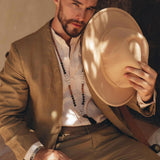 A man sits on a chair wearing an AYA Sacred Wear Brown Linen Blazer Jacket and a white shirt, holding a beige fedora hat in one hand. Beads adorn his neck, and he wears a contemplative expression while sunlight casts shadows on him. The softly lit background adds a warm ambiance.