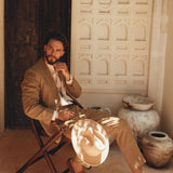 Dressed in a Brown Linen Blazer Jacket for Men by AYA Sacred Wear, a man sits on a wooden chair, cradling a matching hat. In this rustic setting adorned with carved walls and pottery jars, sunlight streams through, casting intricate shadows on the ground.