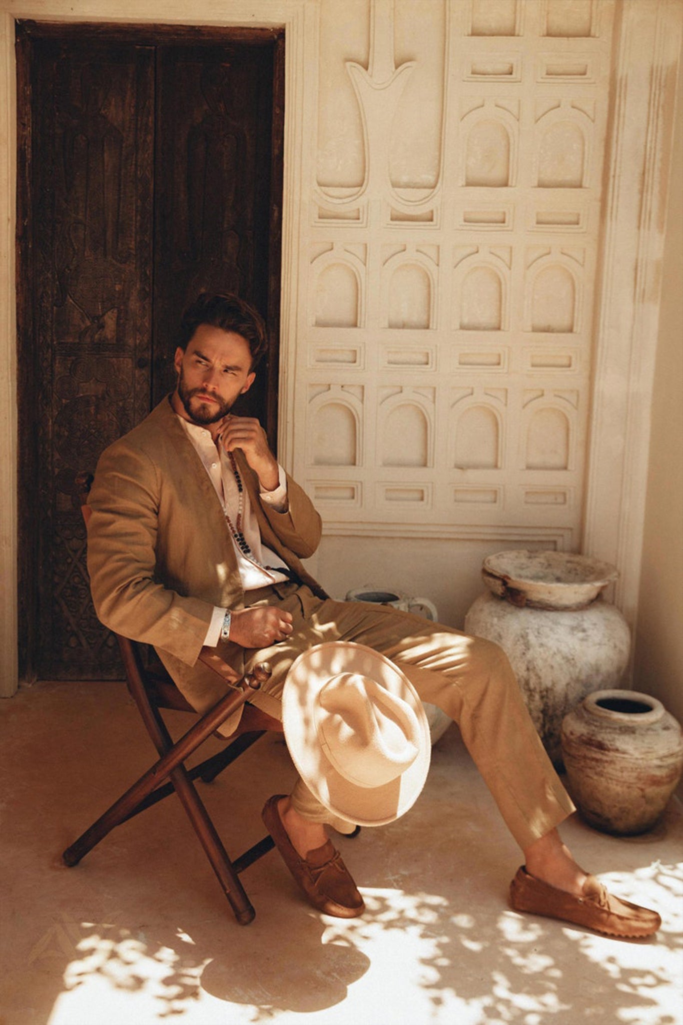 Dressed in a Brown Linen Blazer Jacket for Men by AYA Sacred Wear, a man sits on a wooden chair, cradling a matching hat. In this rustic setting adorned with carved walls and pottery jars, sunlight streams through, casting intricate shadows on the ground.