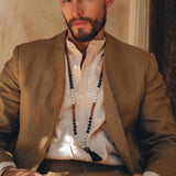 A man with a beard wearing the Brown Linen Blazer Jacket for Men from AYA Sacred Wear and a white shirt sits against a wall. He is adorned with a long beaded necklace. The warm lighting accentuates his relaxed and confident posture.