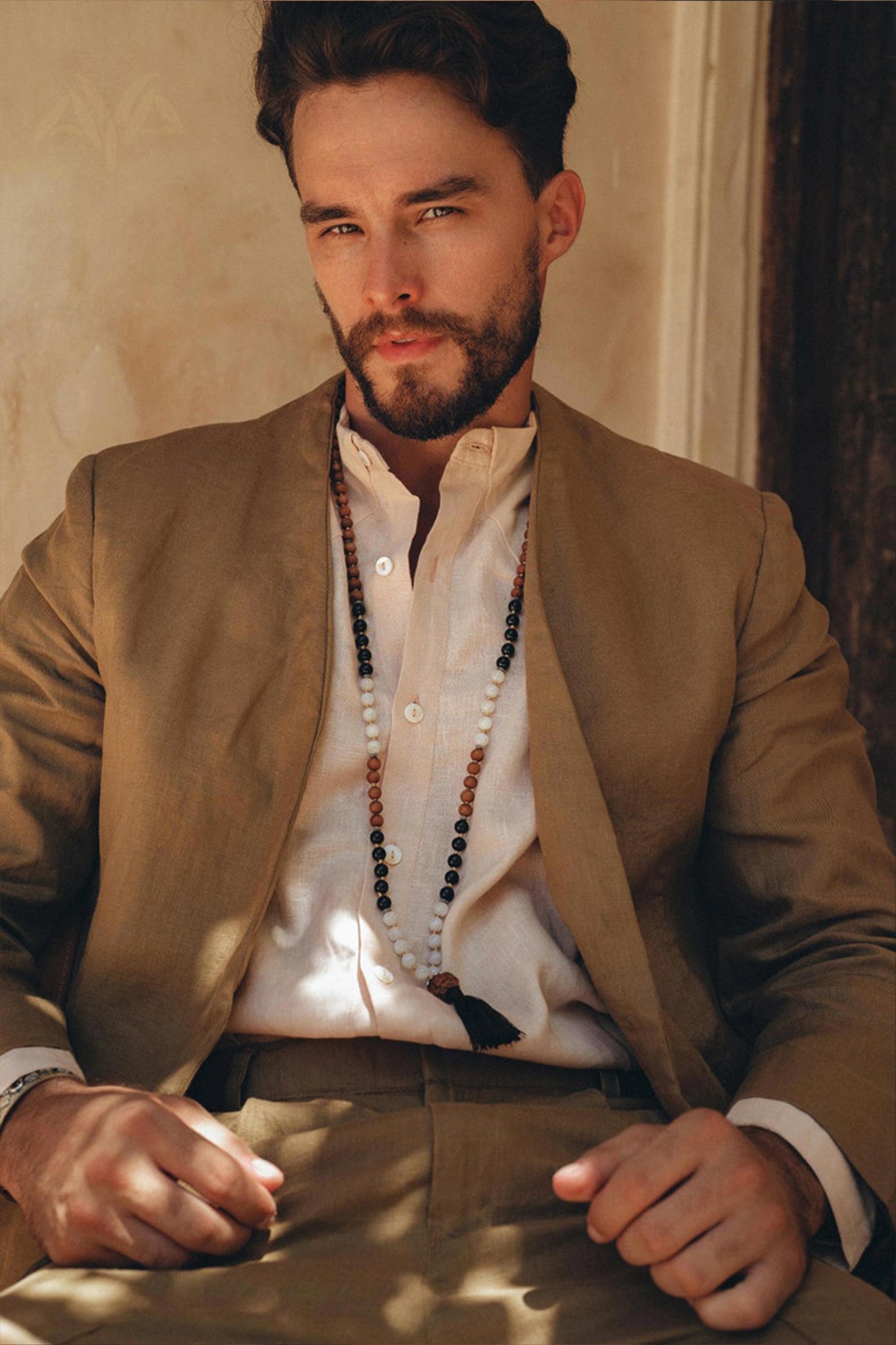 A man with a beard wearing the Brown Linen Blazer Jacket for Men from AYA Sacred Wear and a white shirt sits against a wall. He is adorned with a long beaded necklace. The warm lighting accentuates his relaxed and confident posture.