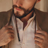 A bearded man is dressed in a light shirt and a wooden bead necklace, holding the lapels of his AYA Sacred Wear Brown Linen Blazer Jacket for Men with both hands as he gazes thoughtfully to the side. Warm lighting casts soft shadows across his face.