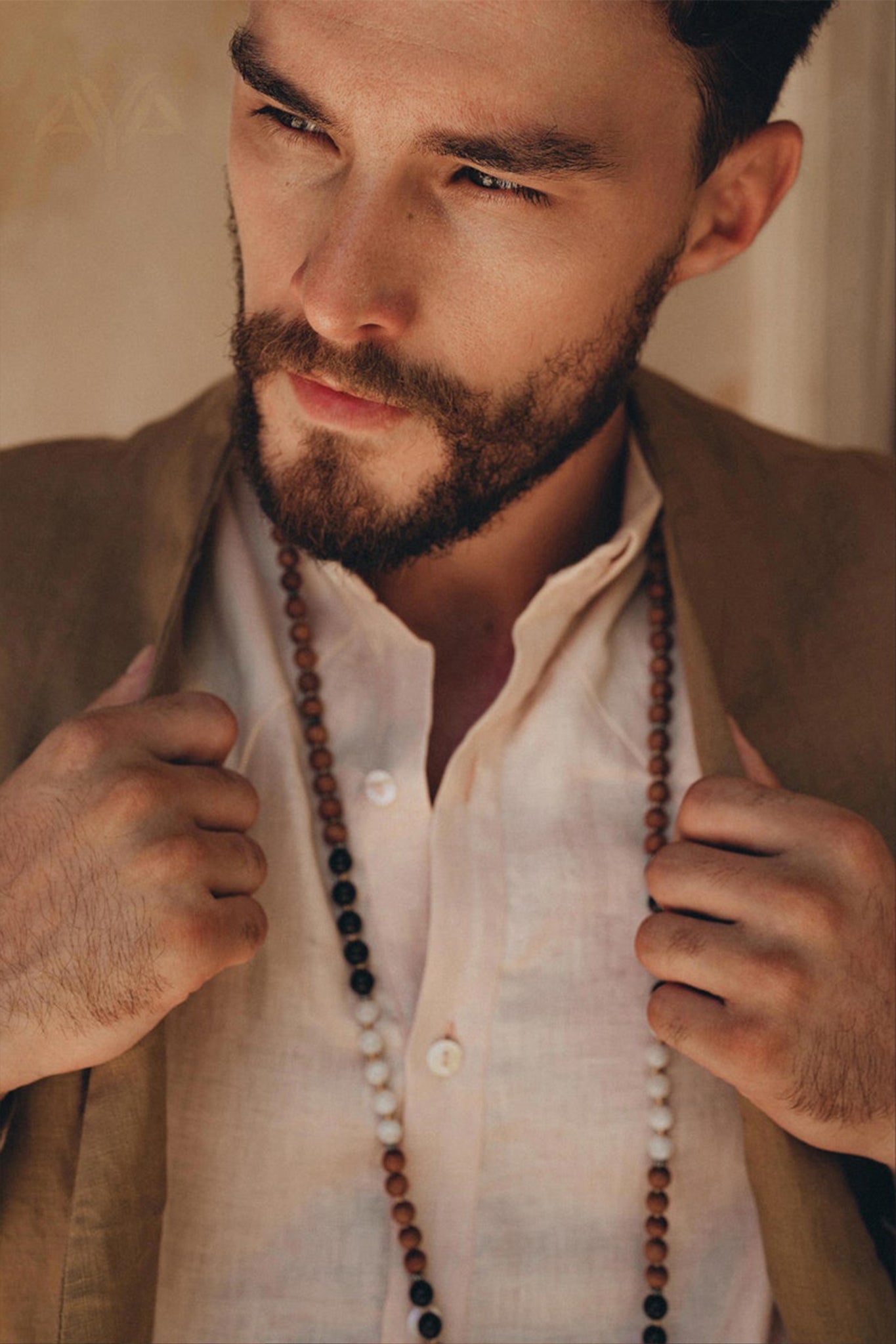 A bearded man is dressed in a light shirt and a wooden bead necklace, holding the lapels of his AYA Sacred Wear Brown Linen Blazer Jacket for Men with both hands as he gazes thoughtfully to the side. Warm lighting casts soft shadows across his face.