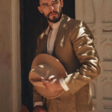 A man dressed in a Brown Linen Blazer Jacket by AYA Sacred Wear stands with a neutral expression, holding a matching hat. The scene features an ornate, light-colored architectural background with intricate designs. His blazer adds an artistic touch to the setting, blending seamlessly into the elegant surroundings.