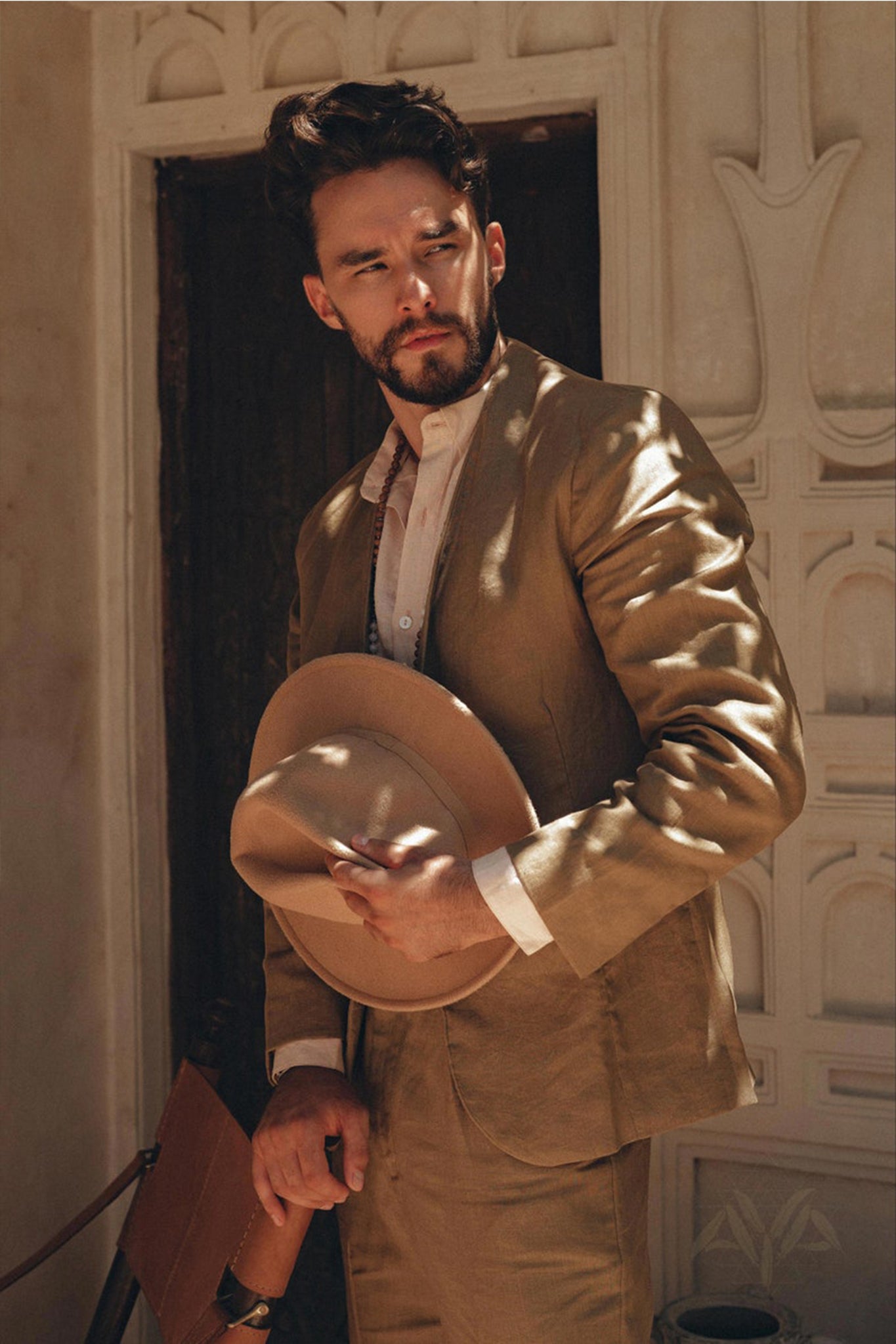 A man dressed in a Brown Linen Blazer Jacket by AYA Sacred Wear stands with a neutral expression, holding a matching hat. The scene features an ornate, light-colored architectural background with intricate designs. His blazer adds an artistic touch to the setting, blending seamlessly into the elegant surroundings.