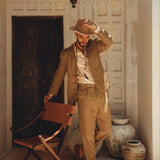 A person dressed in the Brown Linen Blazer Jacket for Men by AYA Sacred Wear stands on a rustic porch, adjusting the brim of their tan hat with one hand while resting the other on a wooden chair. Behind them, a closed wooden door and large clay pots cast shadows in the sunlight.