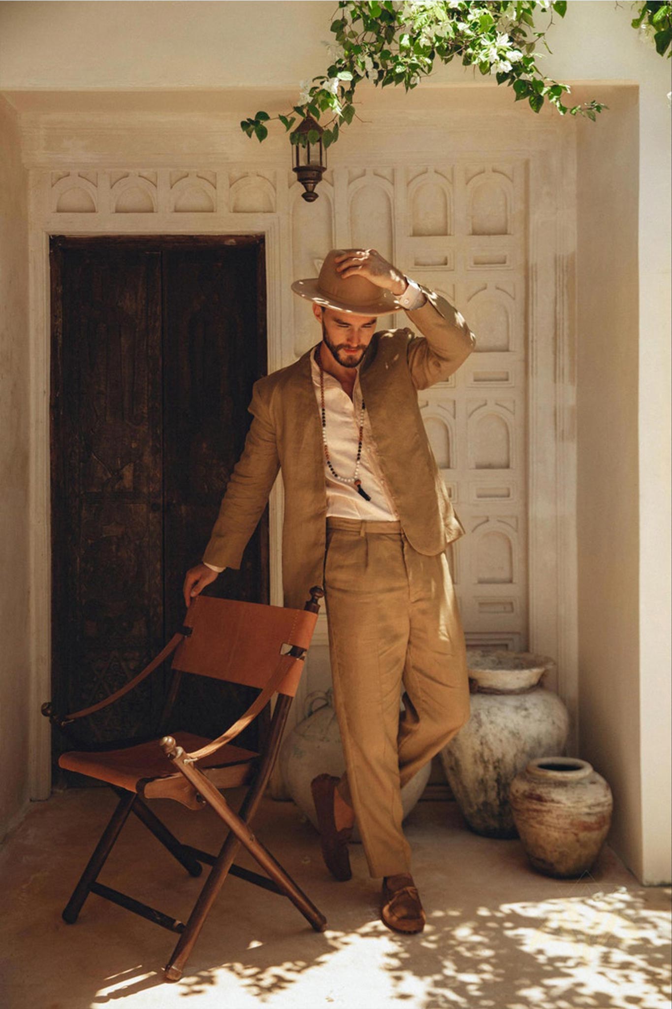 A person dressed in the Brown Linen Blazer Jacket for Men by AYA Sacred Wear stands on a rustic porch, adjusting the brim of their tan hat with one hand while resting the other on a wooden chair. Behind them, a closed wooden door and large clay pots cast shadows in the sunlight.