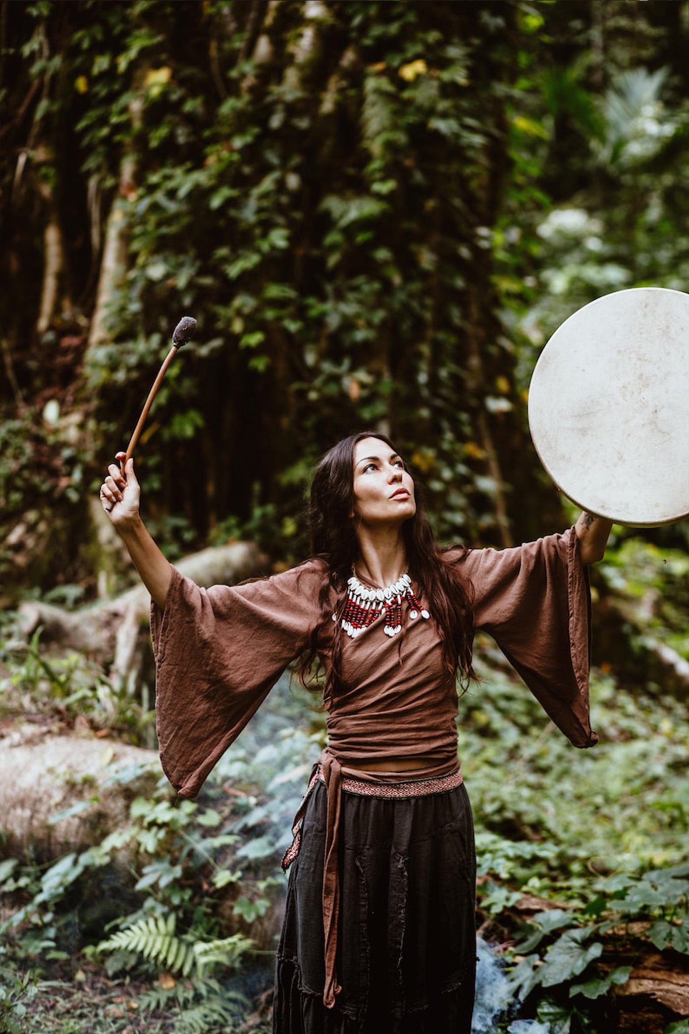 In a lush forest, a person stands focused, wearing the "Chi Wrap Top," a sustainable linen kimono wrap in earthy tones by AYA Sacred Wear. They raise a drum with one hand and hold the stick in the other, surrounded by vibrant greenery and towering tree trunks.