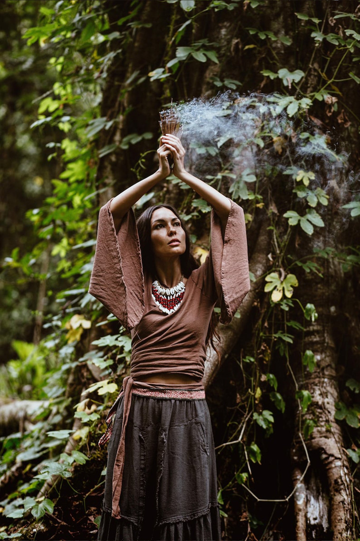 In the heart of a forest, an individual stands with arms raised, holding a bundle of smoking herbs. They are draped in AYA Sacred Wear's handwoven Brown Linen Kimono Wrap "Chi Wrap Top" paired with a flowing skirt, complemented by a beaded necklace. The lush green foliage envelops them, contributing to an earthy and mystical ambiance.