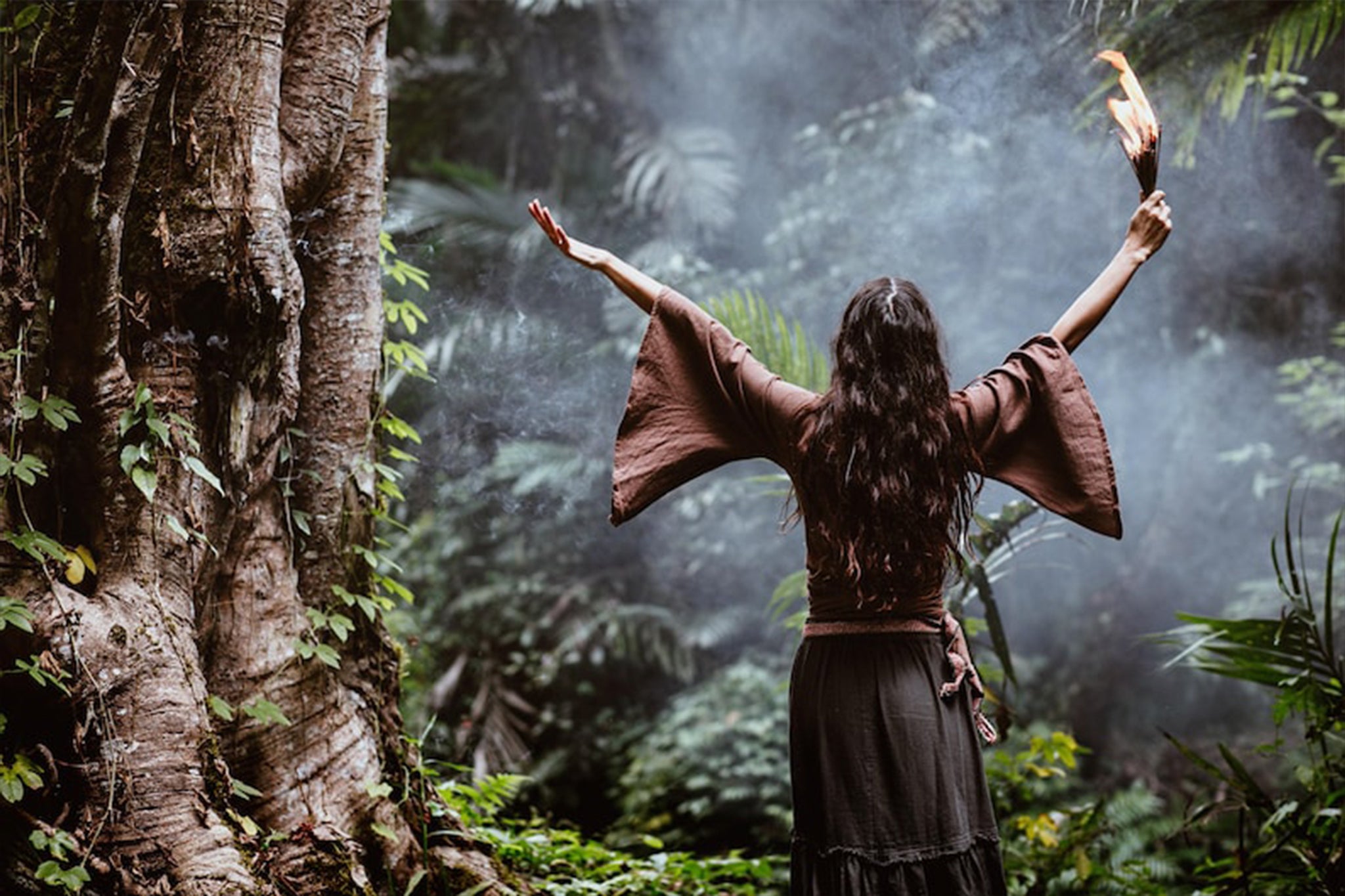 In the midst of a lush forest, an individual stands with arms raised, holding a torch while wearing the earthy-toned "Chi Wrap Top," a brown linen kimono blouse with wide sleeves by AYA Sacred Wear. Smoke swirls around them, and they're surrounded by large trees and dense foliage that add to the mystical atmosphere.