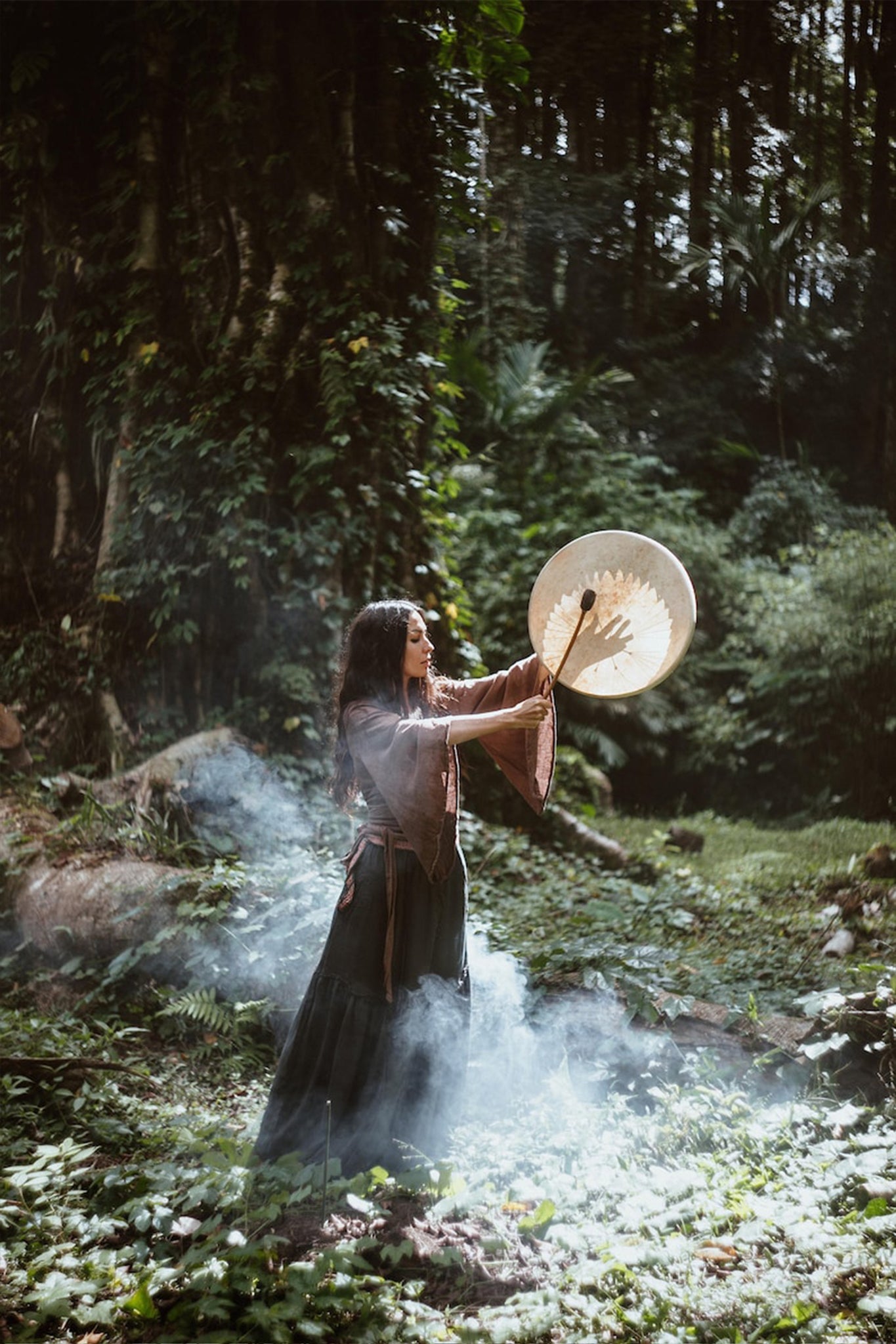 In a serene forest clearing enveloped in mist, a woman stands with a large drum raised towards the sky. She wears the Brown Linen Kimono Wrap "Chi Wrap Top" by AYA Sacred Wear, which flows elegantly around her. Sunlight streams through the tall trees, adding to the mystical atmosphere.