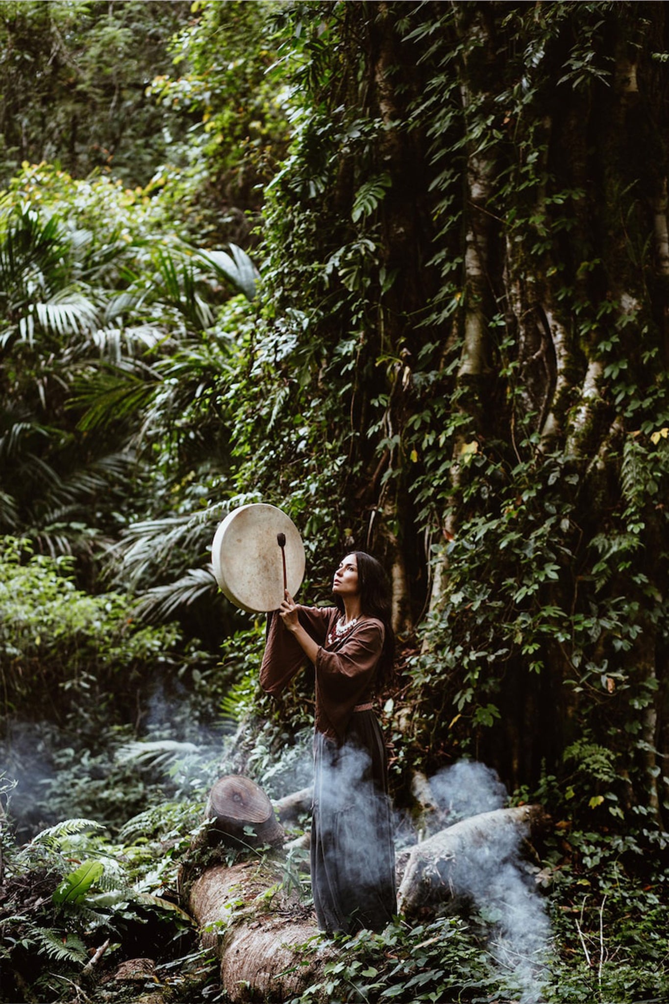 In a lush forest, a person stands holding a large circular drum overhead, dressed in AYA Sacred Wear's sustainable "Chi Wrap Top," a brown linen kimono wrap. Smoke drifts around them, weaving through the dense greenery and handwoven fabric, evoking a mystical and serene atmosphere.