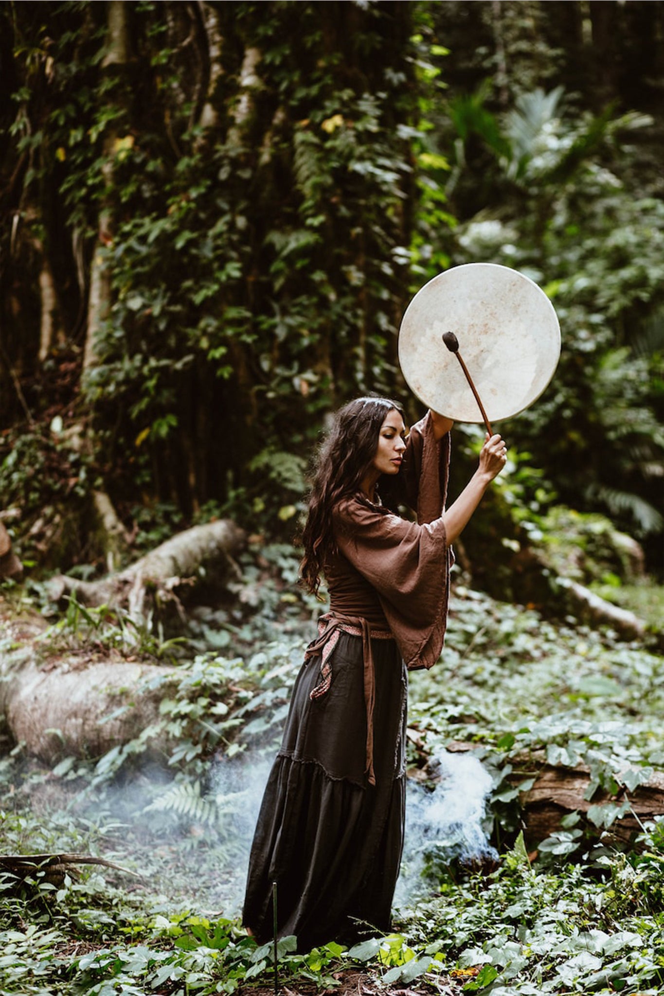 Dressed in the Brown Linen Kimono Wrap "Chi Wrap Top" by AYA Sacred Wear, a person stands amidst a lush forest, lifting a large round drum over their head. Smoke swirls around them, adding to the mystical atmosphere amid the dense, green foliage.