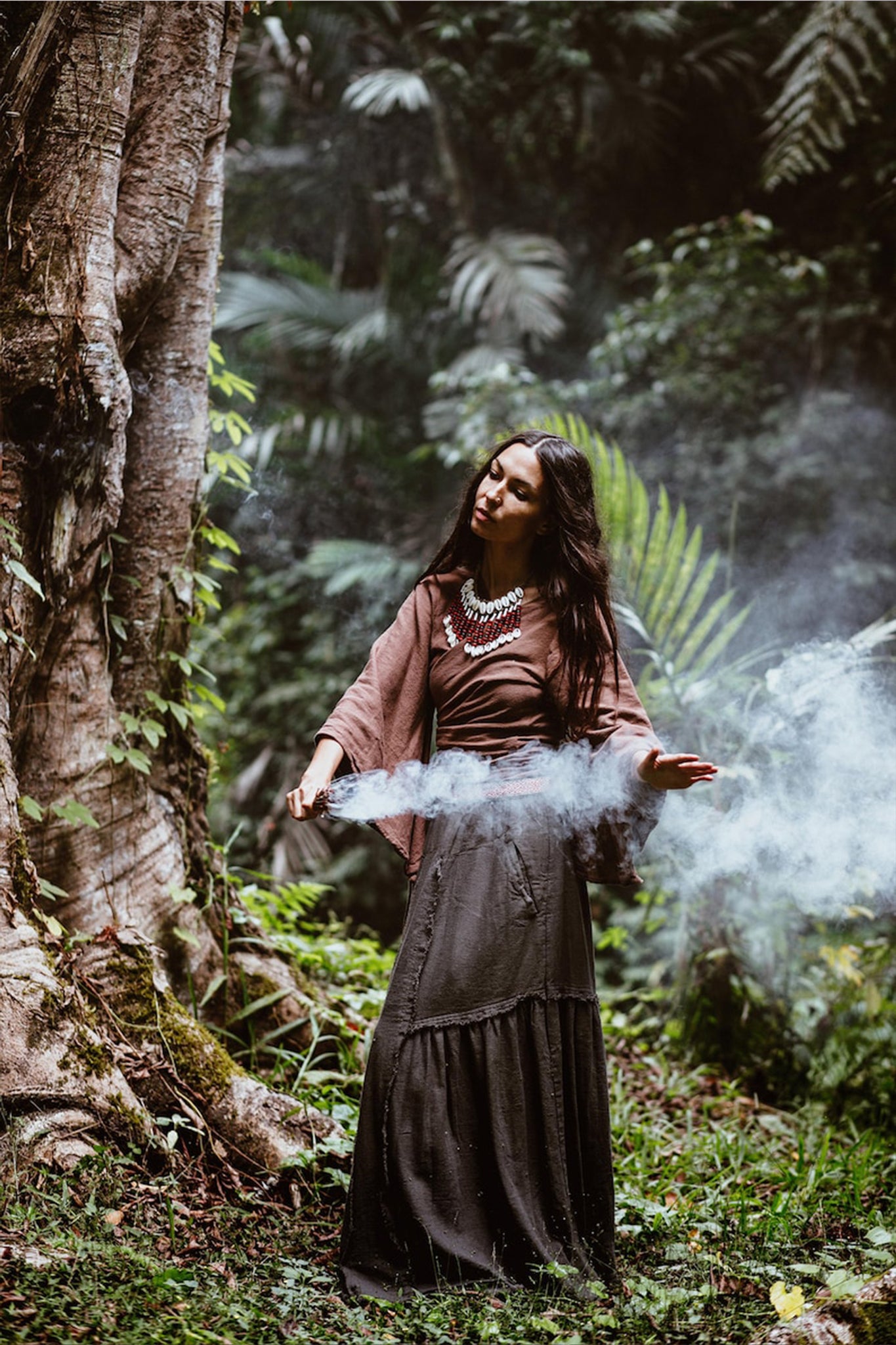 A woman in the serene embrace of a lush forest, surrounded by towering trees and rich greenery, stands gracefully in the Brown Linen Kimono Wrap "Chi Wrap Top" by AYA Sacred Wear. Holding a stick that releases wisps of aromatic smoke, she conjures a mystical atmosphere. Sunlight gently pierces through the leafy canopy above, accentuating the soft texture of her sustainable linen attire.