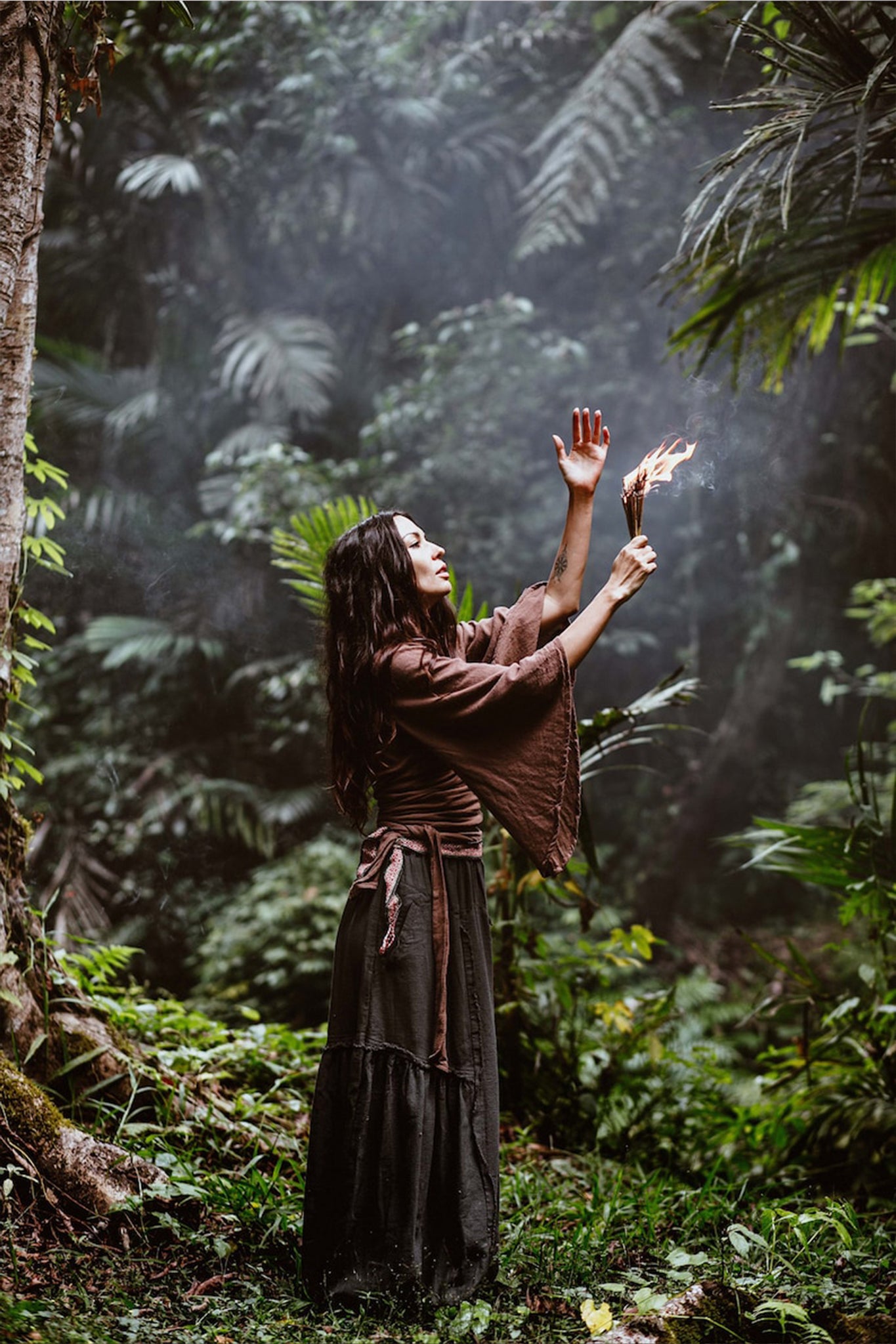 A woman stands in a forest, draped in the Brown Linen Kimono Wrap "Chi Wrap Top" by AYA Sacred Wear, paired with a long handwoven skirt. She holds a burning torch aloft, enveloped by lush greenery and mist that evoke a mystical atmosphere.