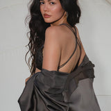 A woman with long, wavy dark hair gazes over her shoulder, revealing the AYA Sacred Wear Butterfly Silk Bra in Asphalt Gray. Draped in a handcrafted Bali silk shawl, she stands before a soft, textured background for a minimal, elegant look.