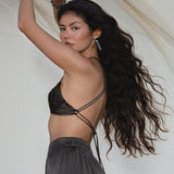 A woman with long, wavy dark hair poses sideways, wearing the AYA Sacred Wear Butterfly Silk Bra in Asphalt Gray. She holds a light fabric backdrop behind her as lighting accentuates her smooth skin and flowing hair.