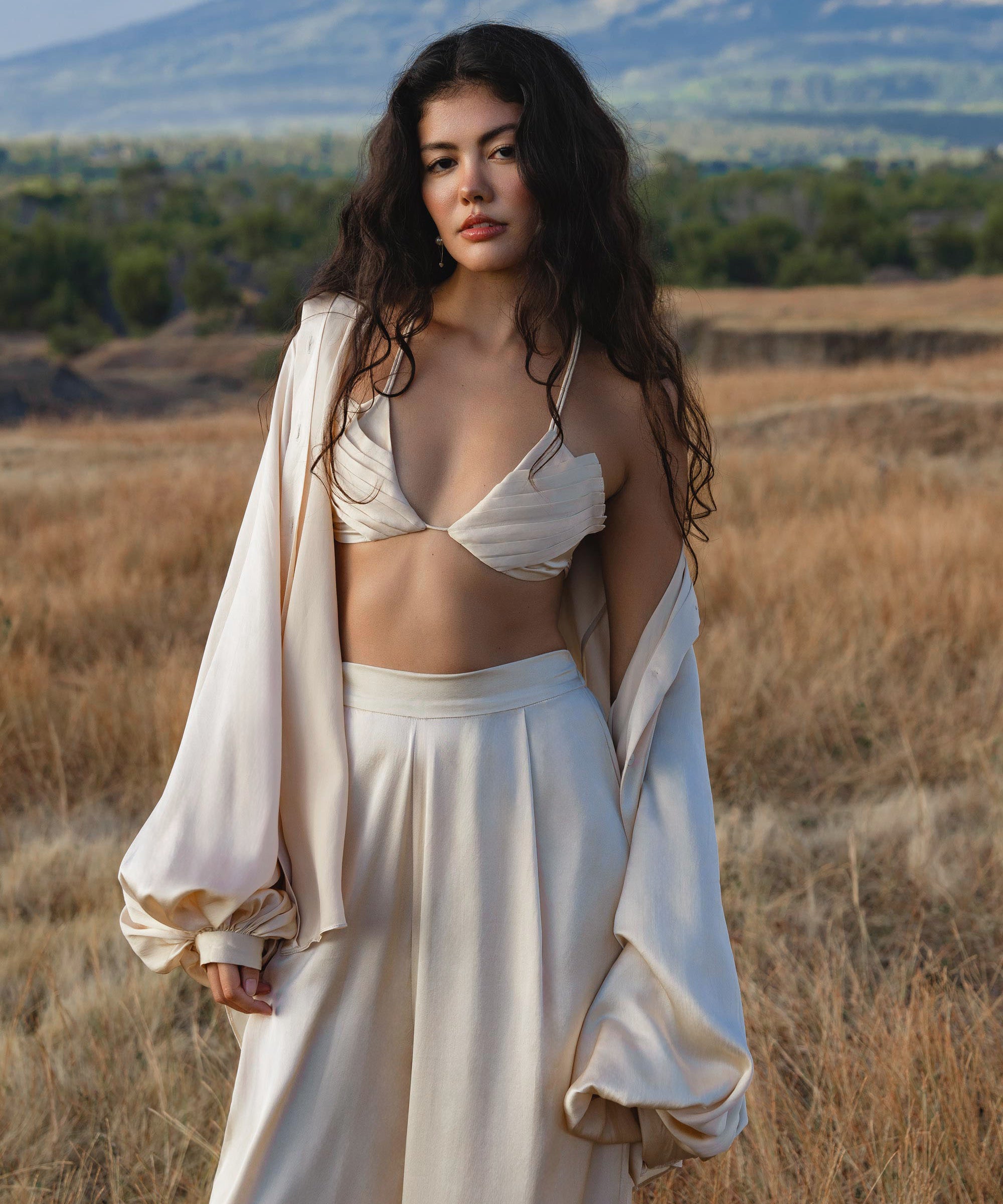 A woman with long dark wavy hair stands in a dry grassy field wearing the Butterfly Silk Bra Cream – Pure Silk Bralette Handmade by AYA Sacred Wear, paired with wide-legged pants and a draped jacket. Mountains and clouds appear behind her as she gazes calmly at the camera.