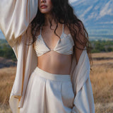 A woman with long, wavy dark hair poses outdoors in a flowing outfit and the Butterfly Silk Bra Cream – Pure Silk Bralette Handmade by AYA Sacred Wear, standing in a grassy field with mountains and blue sky behind her.