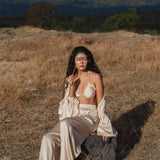 A woman with long dark hair sits on a rock in a dry, grassy field wearing the Butterfly Silk Bra Cream – a pure silk bralette handmade by AYA Sacred Wear. Green trees and a cloud-capped mountain rise behind her under sunny skies.