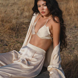 A woman with long, wavy black hair sits on a rock in a dry, grassy field wearing the Butterfly Silk Bra Cream – Pure Silk Bralette Handmade by AYA Sacred Wear, paired with wide-leg pants and an open jacket, gazing confidently at the camera.