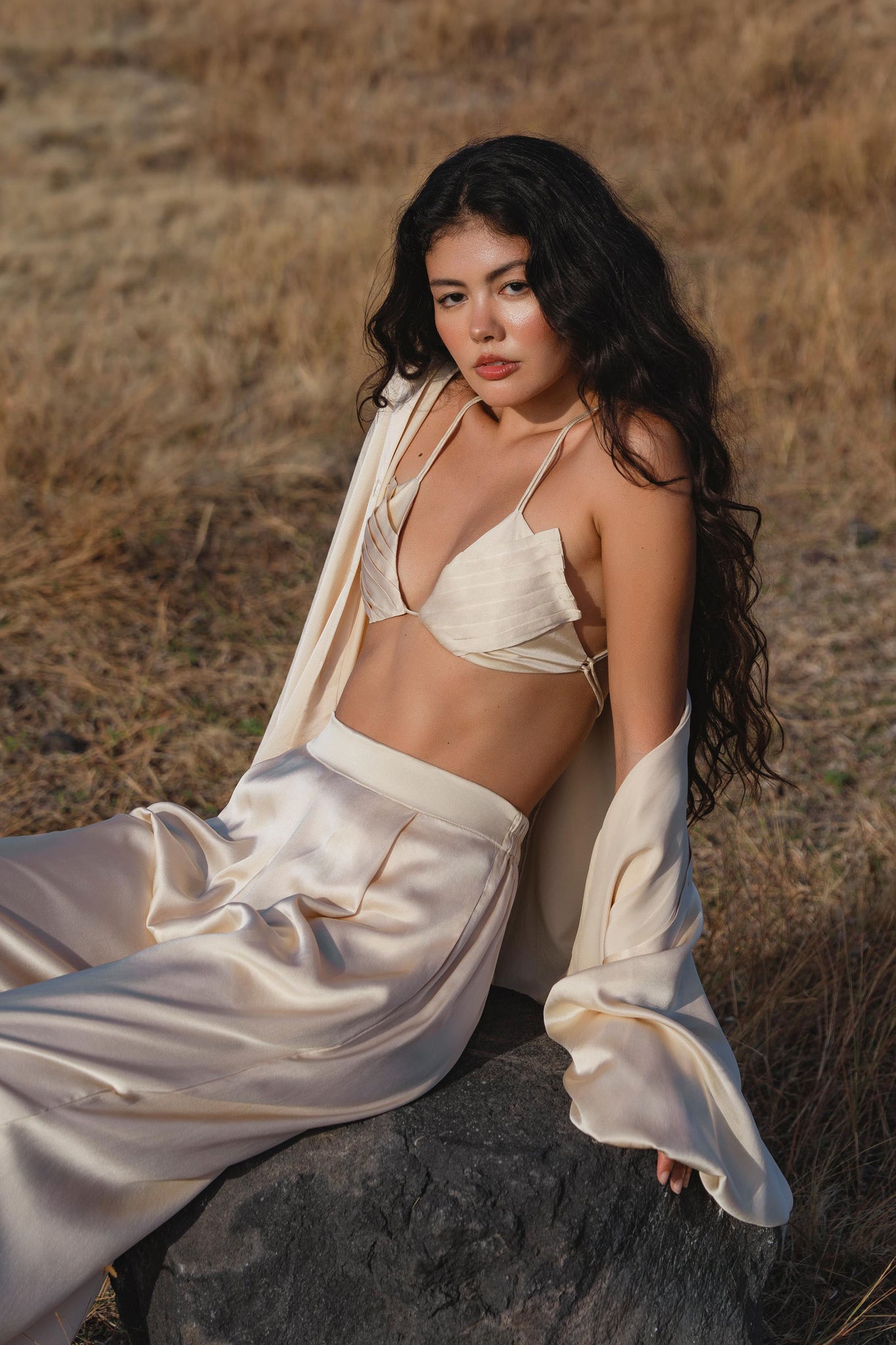 A woman with long, wavy black hair sits on a rock in a dry, grassy field wearing the Butterfly Silk Bra Cream – Pure Silk Bralette Handmade by AYA Sacred Wear, paired with wide-leg pants and an open jacket, gazing confidently at the camera.