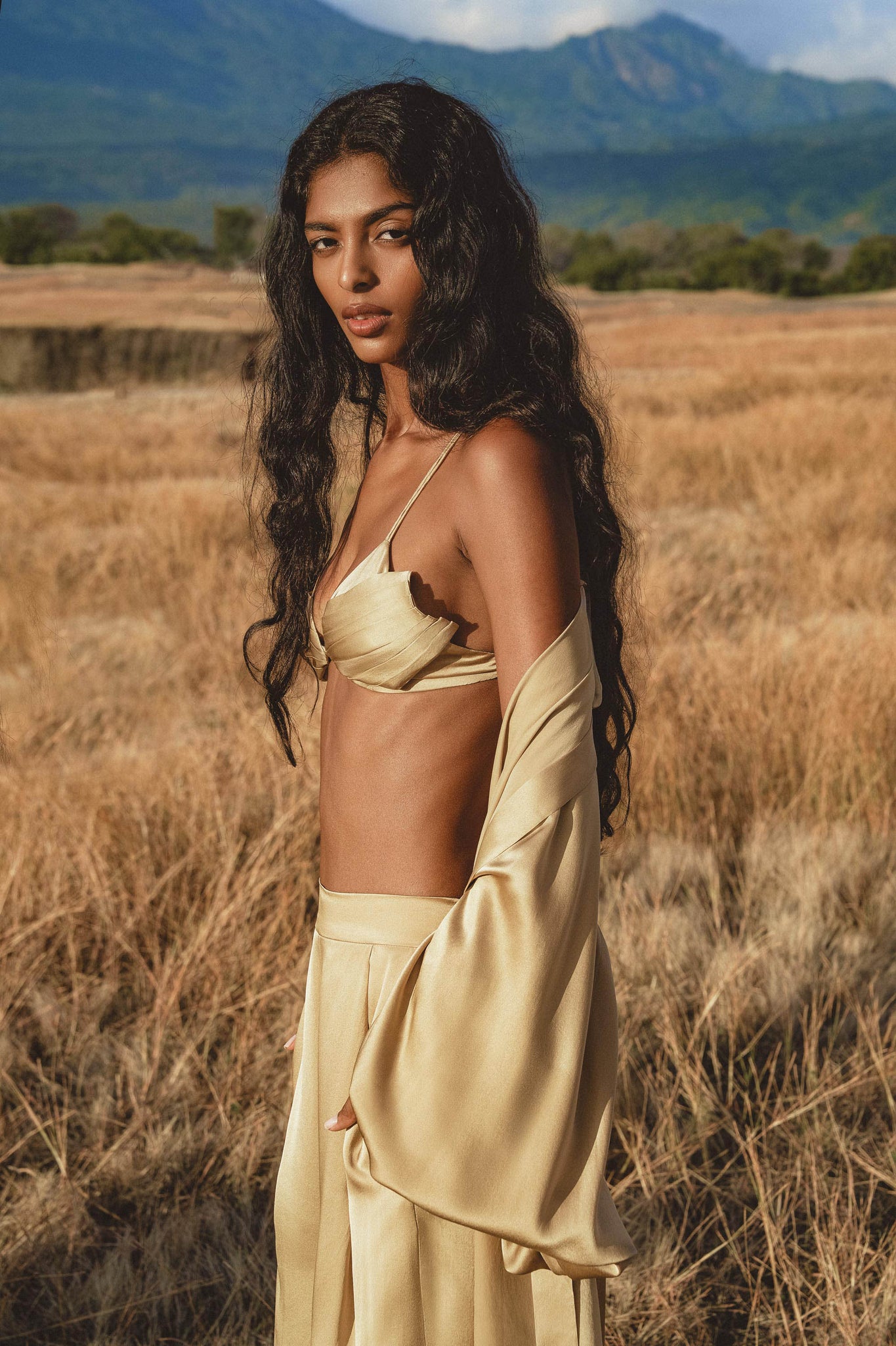 A woman with long wavy dark hair stands in a sunlit field wearing the Butterfly Silk Bra Inka Gold—an AYA Sacred Wear pure silk bralette handmade in Bali—as distant green hills and blue mountains frame the partly cloudy sky.