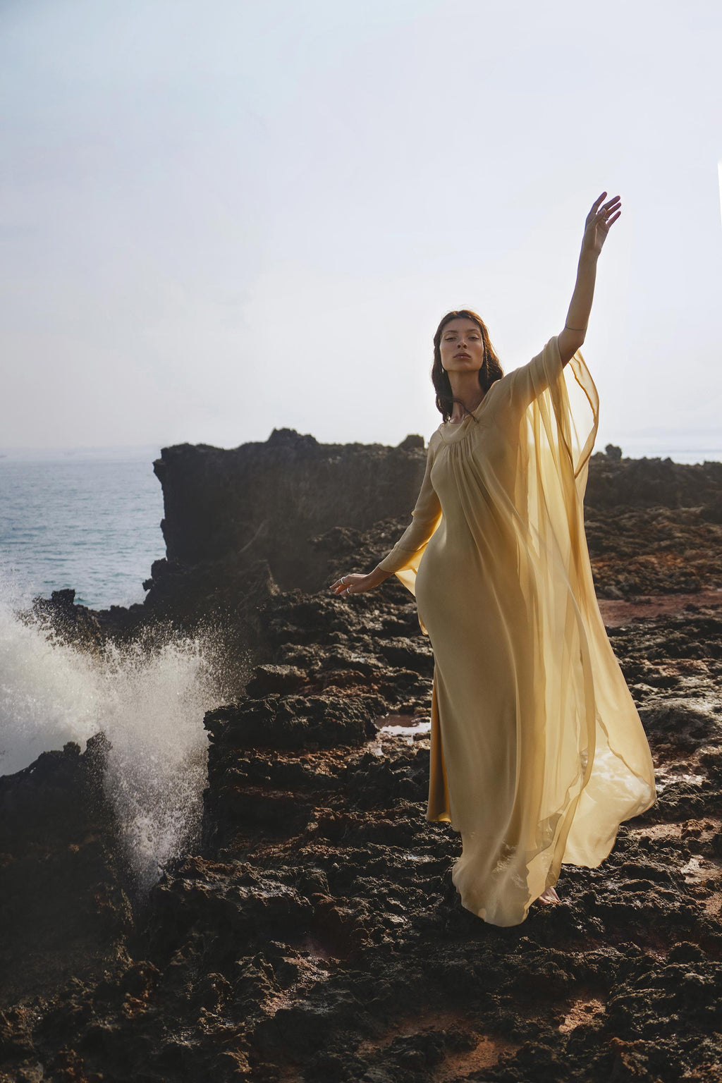 A woman stands gracefully on rocky seaside cliffs in the Mahadevi Dress Inka Gold by AYA Sacred Wear—a pure silk chiffon goddess gown, effortlessly elegant and handcrafted in Bali as ocean waves crash nearby under a hazy sky.