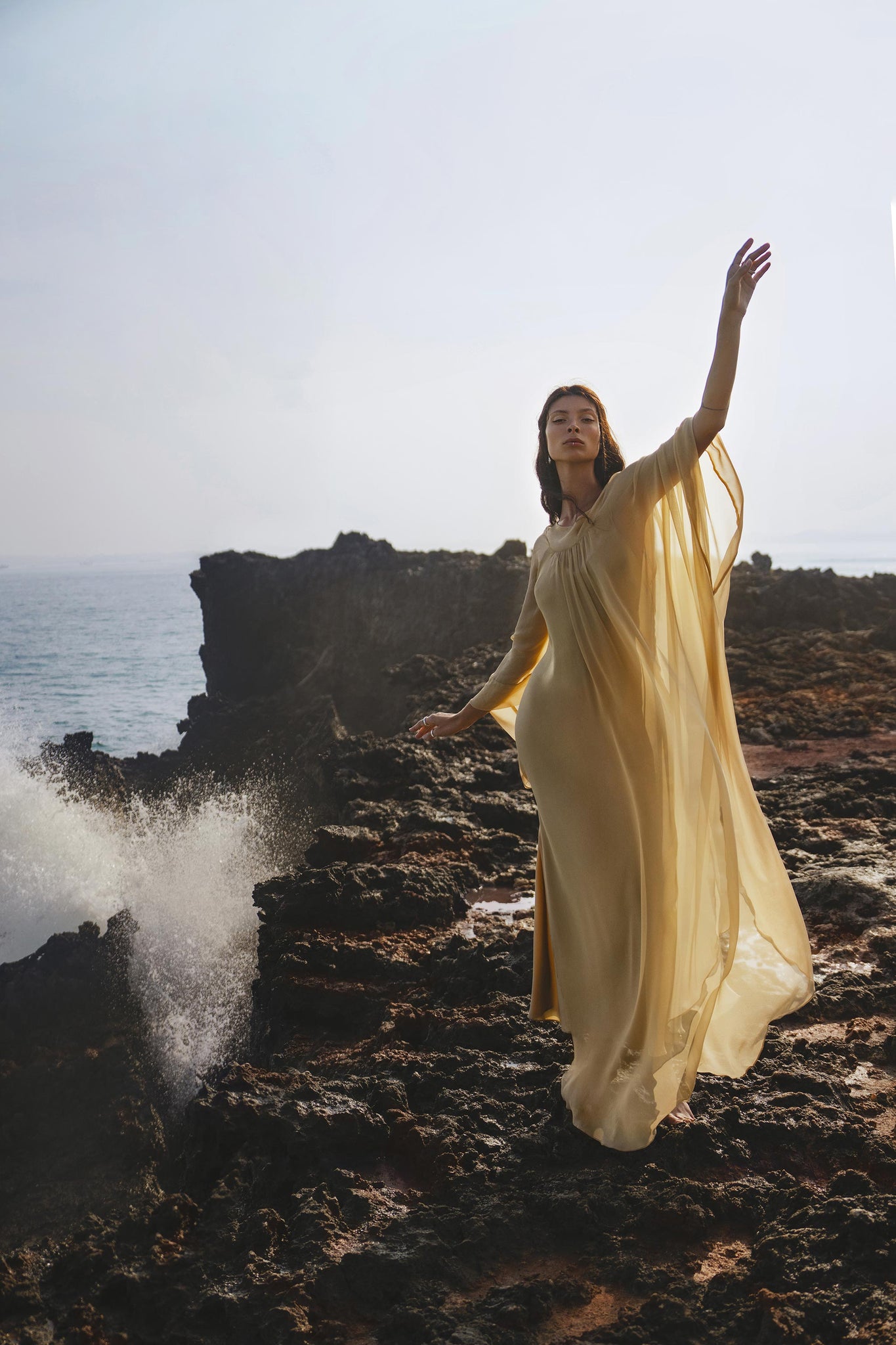 A woman stands gracefully on rocky seaside cliffs in the Mahadevi Dress Inka Gold by AYA Sacred Wear—a pure silk chiffon goddess gown, effortlessly elegant and handcrafted in Bali as ocean waves crash nearby under a hazy sky.