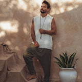 A man stands barefoot on draped fabric, wearing a sleeveless white top and Dark Green Handwoven Cotton Pants from AYA Sacred Wear, crafted from organic cotton. He holds a beaded necklace, with a large potted plant beside him. Sunlit shadows create a dappled effect on the wall behind him.