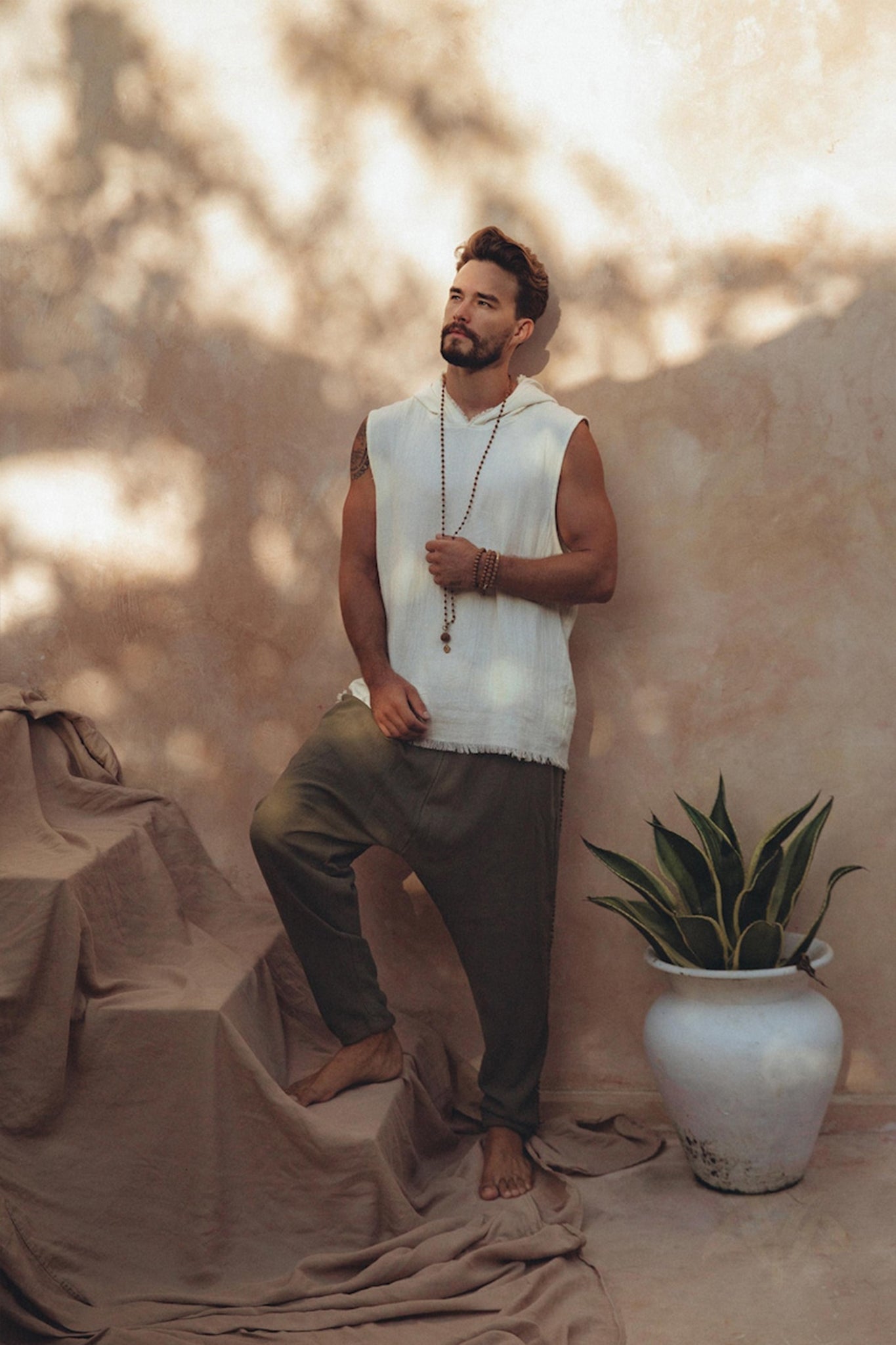 A man stands barefoot on draped fabric, wearing a sleeveless white top and Dark Green Handwoven Cotton Pants from AYA Sacred Wear, crafted from organic cotton. He holds a beaded necklace, with a large potted plant beside him. Sunlit shadows create a dappled effect on the wall behind him.