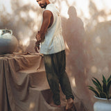 A man poses barefoot on a draped cloth surface, wearing a sleeveless white top and Dark Green Handwoven Cotton Pants for Men by AYA Sacred Wear, crafted from botanically dyed fabric. Shadows and soft light create patterns on the neutral-toned wall and decor, including a large urn and a potted plant.
