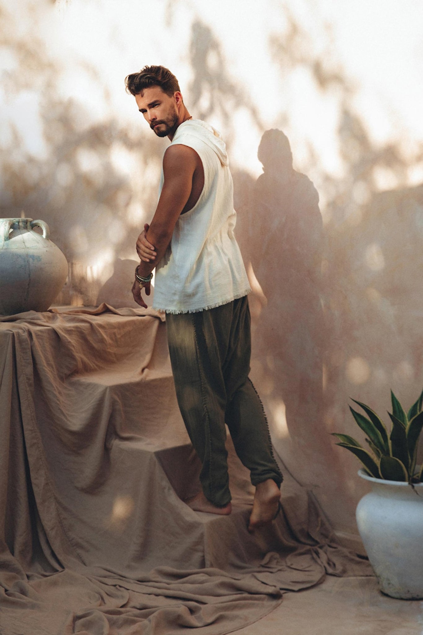 A man poses barefoot on a draped cloth surface, wearing a sleeveless white top and Dark Green Handwoven Cotton Pants for Men by AYA Sacred Wear, crafted from botanically dyed fabric. Shadows and soft light create patterns on the neutral-toned wall and decor, including a large urn and a potted plant.