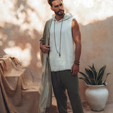 A bearded man stands barefoot on a beige floor, wearing a sleeveless white top and AYA Sacred Wear's Dark Green Handwoven Cotton Pants for Men. He holds a long, handwoven organic cotton scarf and dons a beaded necklace. A potted plant is by his side, with soft shadows enhancing the serene atmosphere.
