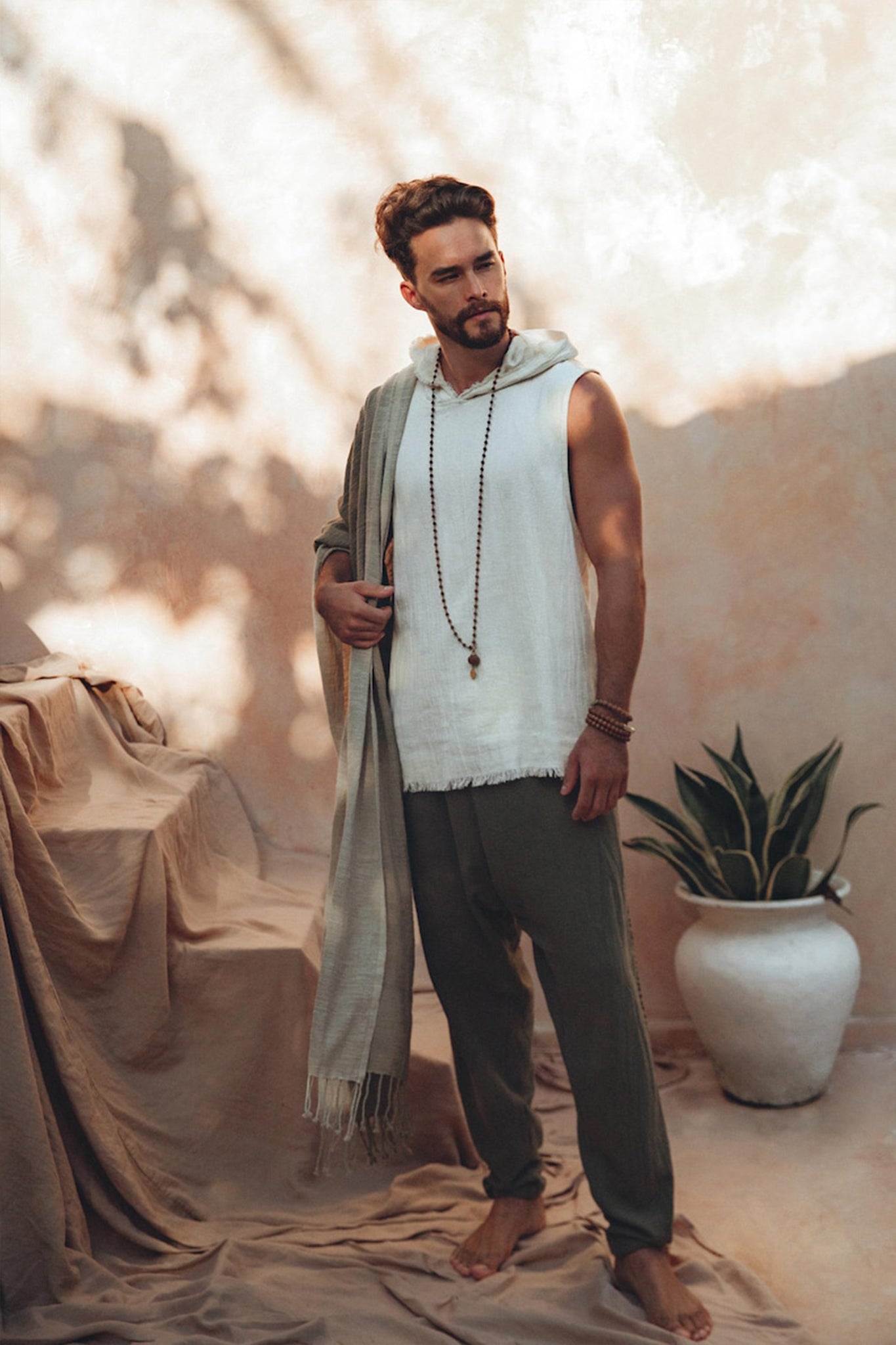 A bearded man stands barefoot on a beige floor, wearing a sleeveless white top and AYA Sacred Wear's Dark Green Handwoven Cotton Pants for Men. He holds a long, handwoven organic cotton scarf and dons a beaded necklace. A potted plant is by his side, with soft shadows enhancing the serene atmosphere.