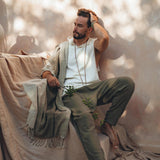 A bearded man sits barefoot on textured fabric, with dappled sunlight creating patterns on the wall. He is dressed in a white shirt, Dark Green Handwoven Cotton Pants for Men from AYA Sacred Wear, and a shawl, holding a leafy branch. His pose is contemplative, with one hand resting on his head.