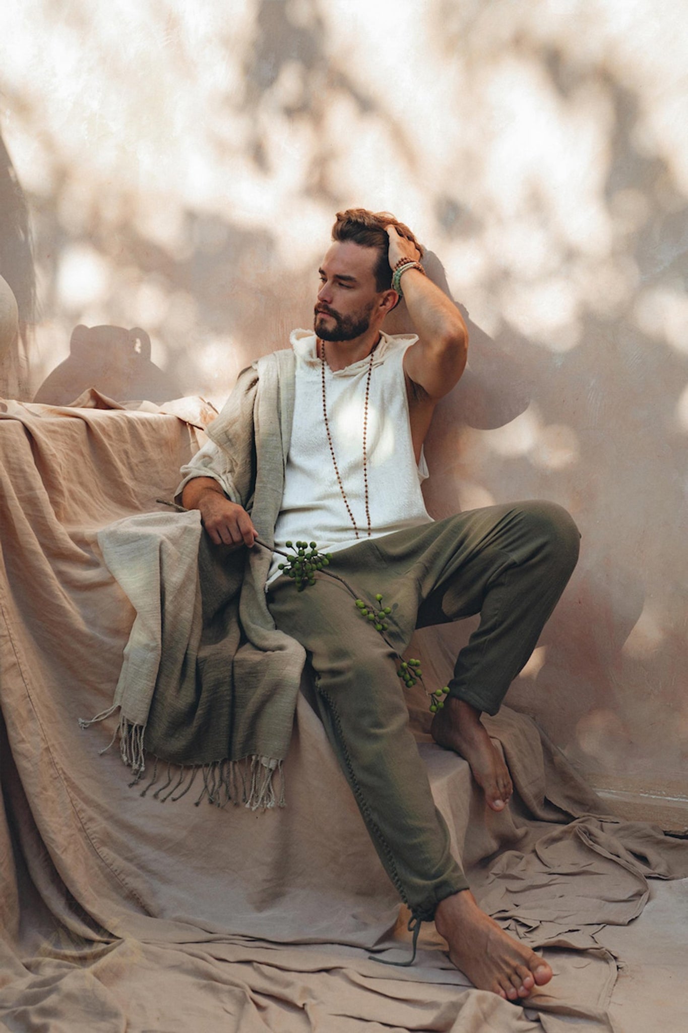 A bearded man sits barefoot on textured fabric, with dappled sunlight creating patterns on the wall. He is dressed in a white shirt, Dark Green Handwoven Cotton Pants for Men from AYA Sacred Wear, and a shawl, holding a leafy branch. His pose is contemplative, with one hand resting on his head.