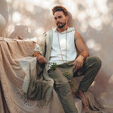 A man with a beard sits barefoot on a draped sofa in a relaxed pose, wearing a white sleeveless top and AYA Sacred Wear's Dark Green Handwoven Cotton Pants for Men, complemented by a long beaded necklace. All are crafted from handwoven organic cotton. Natural light casts leafy shadows on the neutral-toned background.