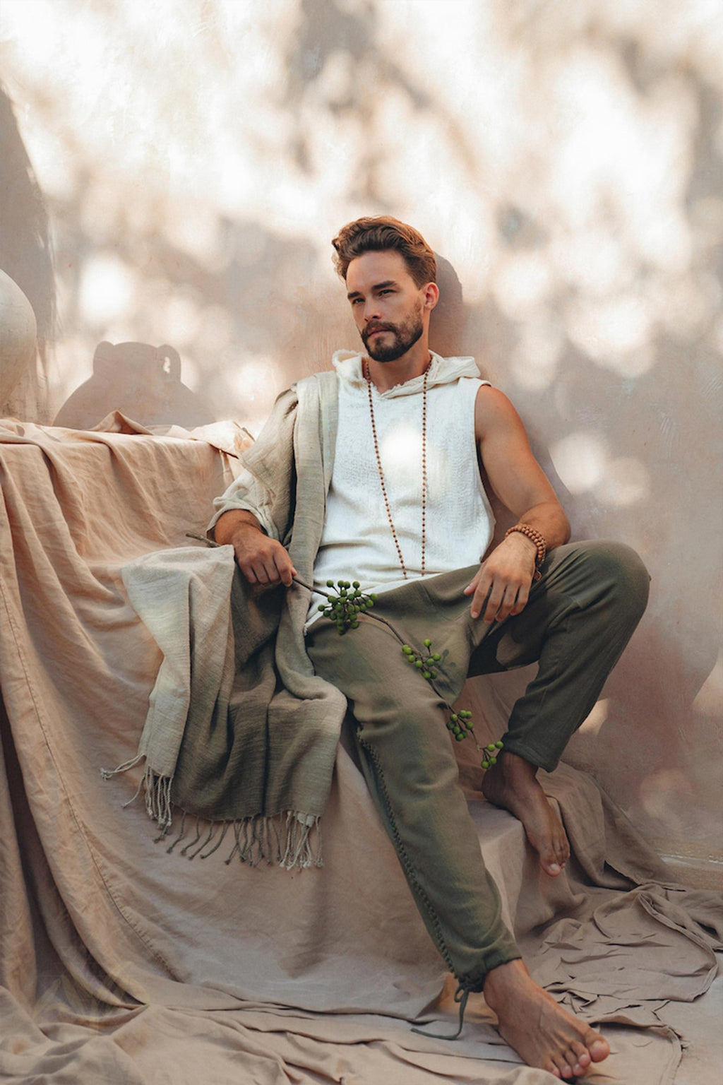 A man with a beard sits barefoot on a draped sofa in a relaxed pose, wearing a white sleeveless top and AYA Sacred Wear's Dark Green Handwoven Cotton Pants for Men, complemented by a long beaded necklace. All are crafted from handwoven organic cotton. Natural light casts leafy shadows on the neutral-toned background.